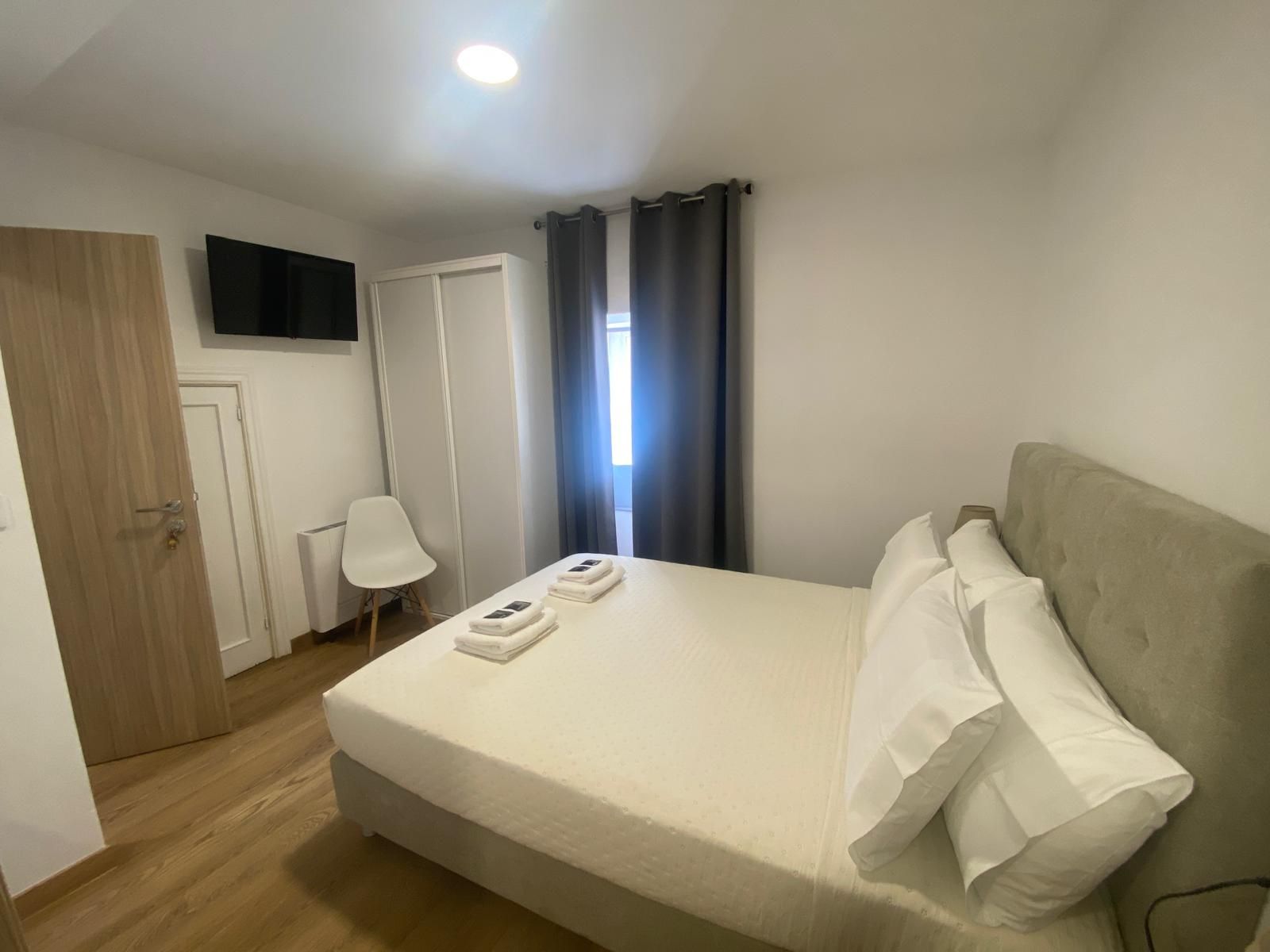 Habitación pequeña y moderna con cama, armario, escritorio, silla, televisor de pared y cortinas grises.