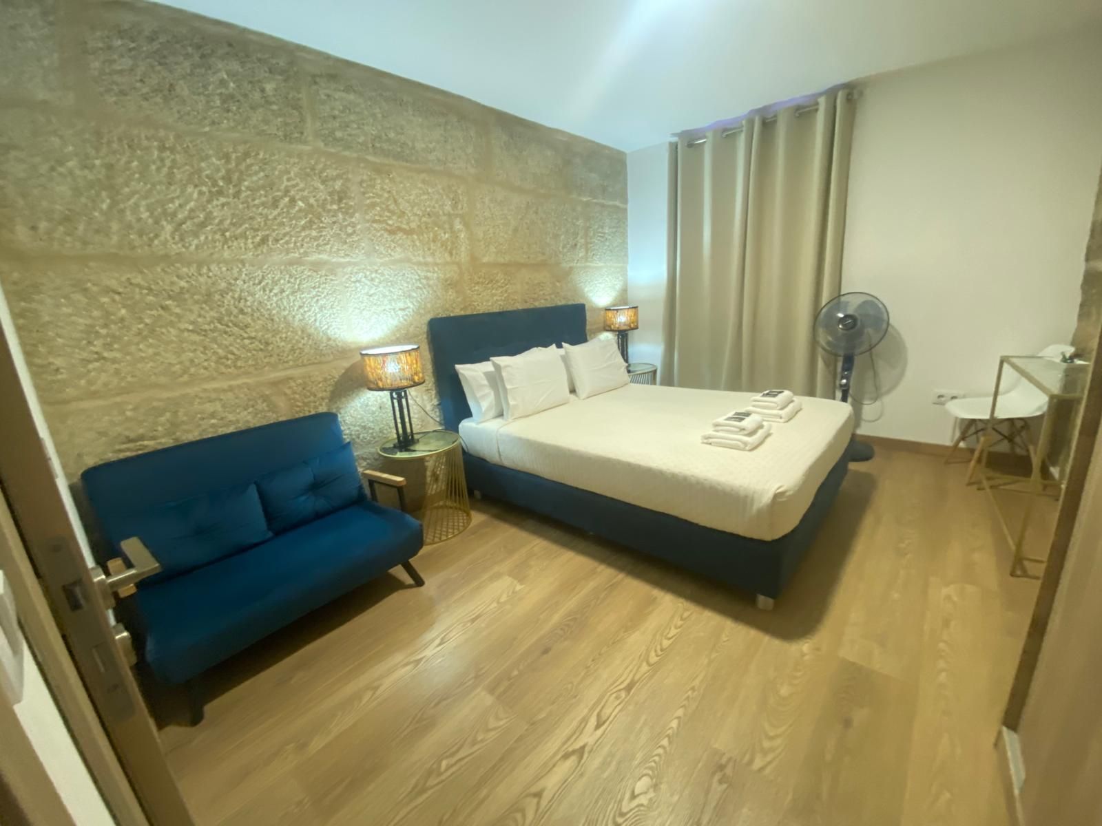 Habitación de hotel con cama king size, sofá azul, lámparas, ventilador y suelo de madera.