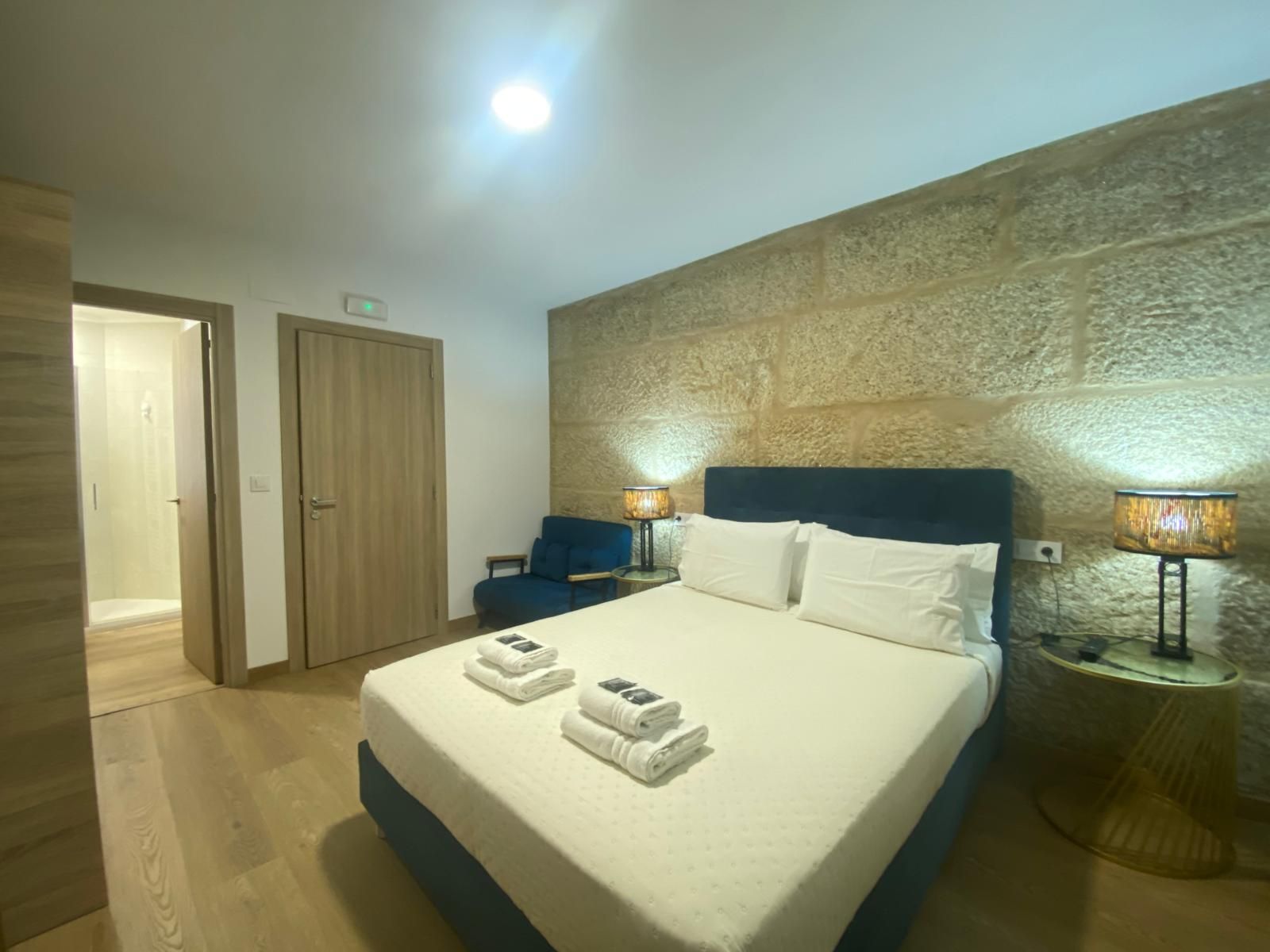 Habitación de hotel luminosa con cama king size, sillas azules, suelo de madera y pared decorativa con textura.