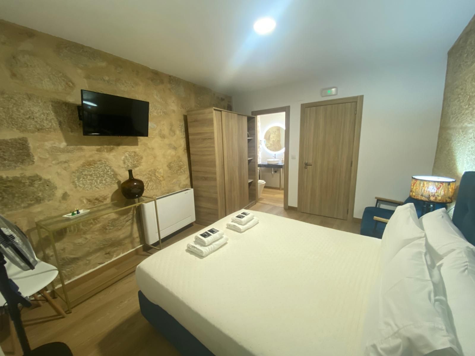 Habitación de hotel moderna con pared decorativa de piedra, cama blanca, televisor y puertas de madera cerca de una ventana.