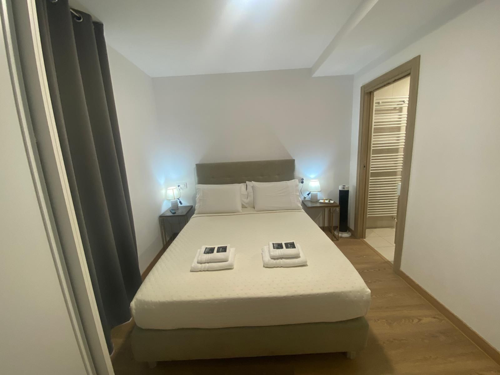 Habitación ordenada con cama doble, cortinas grises, lámparas de noche y una puerta abierta que da a un baño.