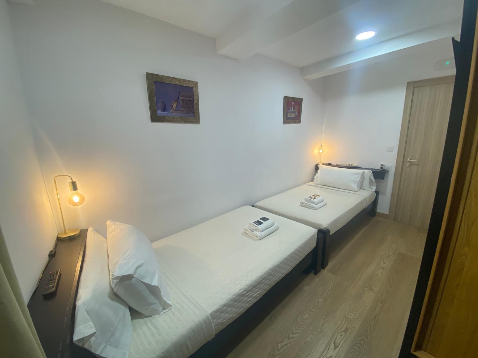 Habitación de hotel pequeña con dos camas individuales, lámparas cálidas, cuadros enmarcados y una puerta cerrada.
