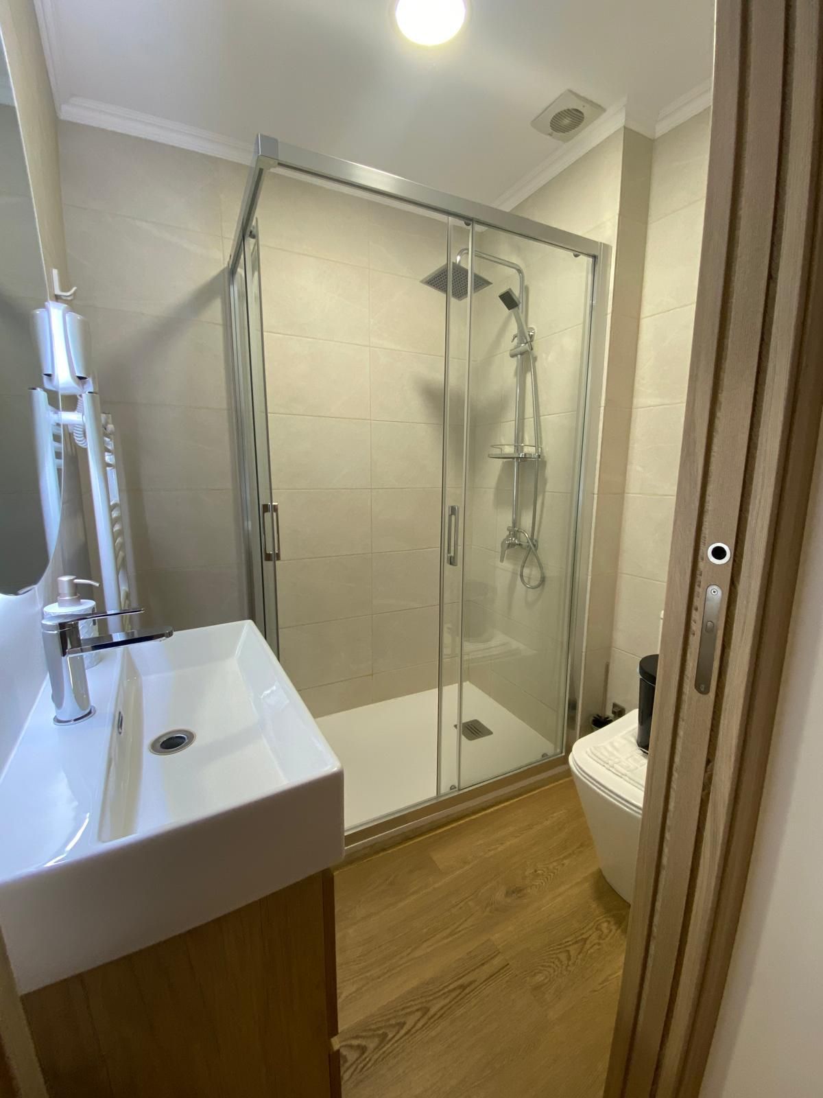 Baño pequeño y moderno con ducha de cristal, lavabo blanco e inodoro sobre suelo de imitación madera.