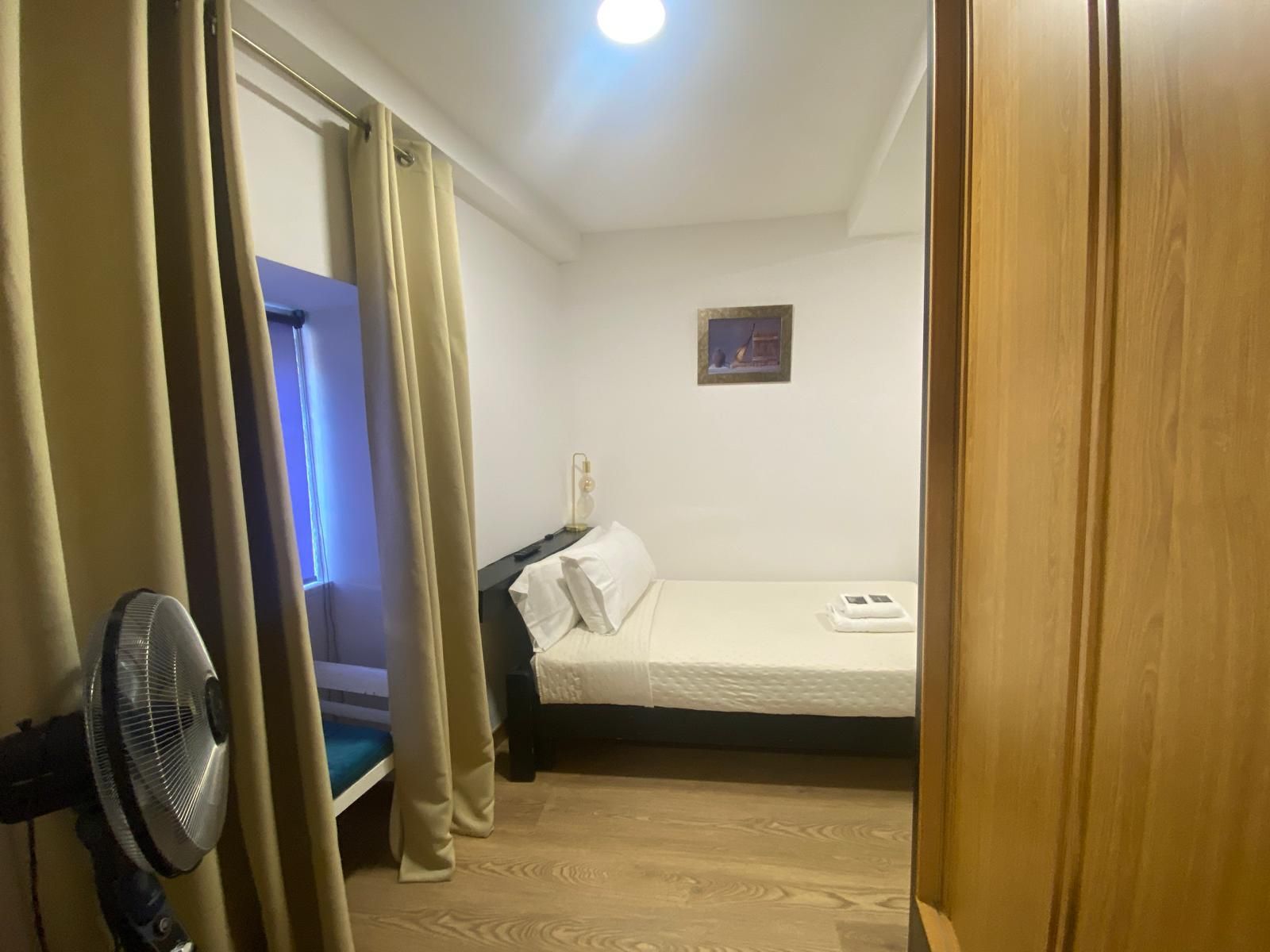 Habitación pequeña con cama, cortinas, ventilador y una puerta iluminada con luz azul.