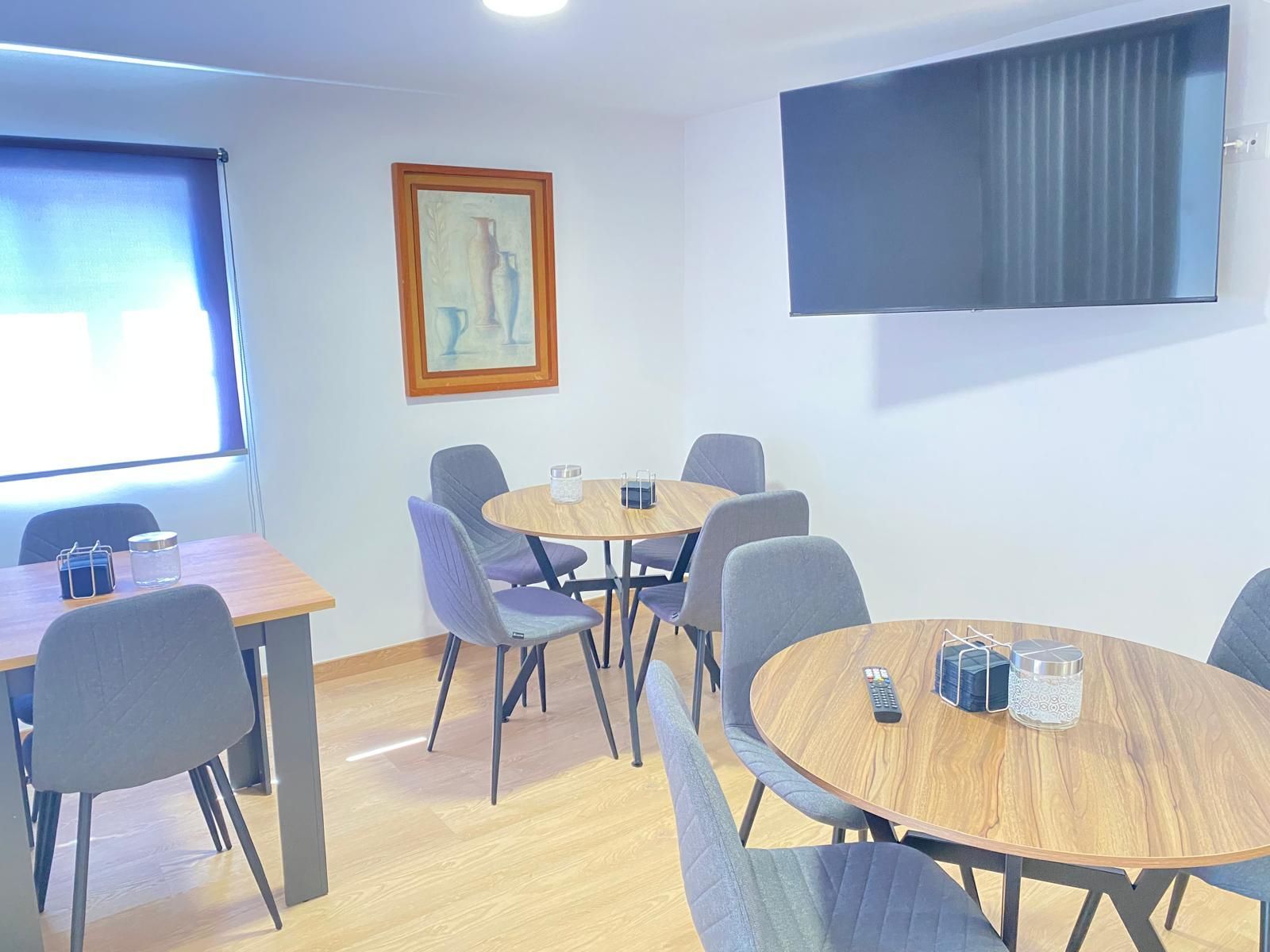 Pequeño comedor con mesas redondas de madera, sillas grises y persianas azules junto a ventanas luminosas.