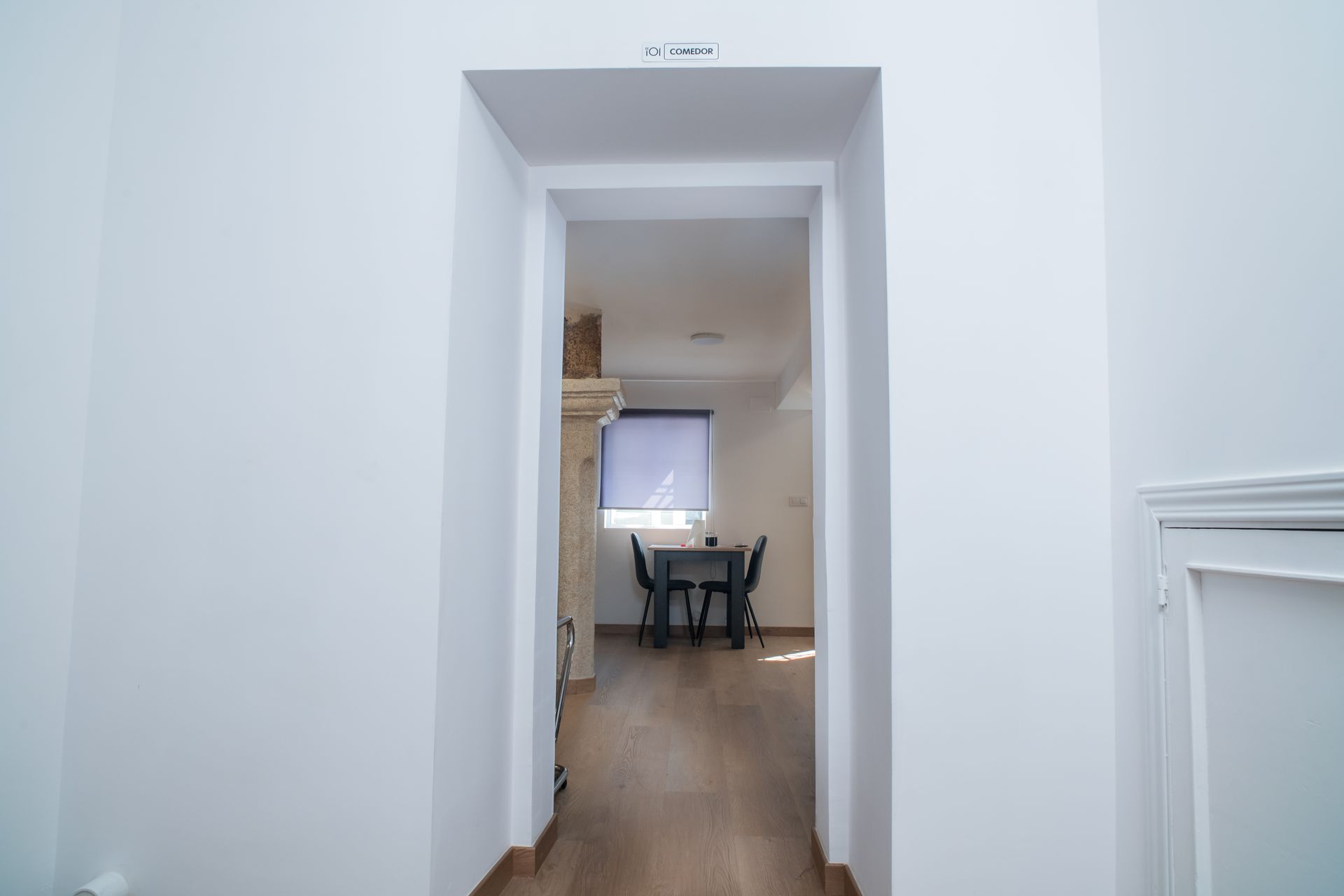 Pasillo blanco que enmarca un comedor con una mesa de madera, sillas y una ventana al fondo.