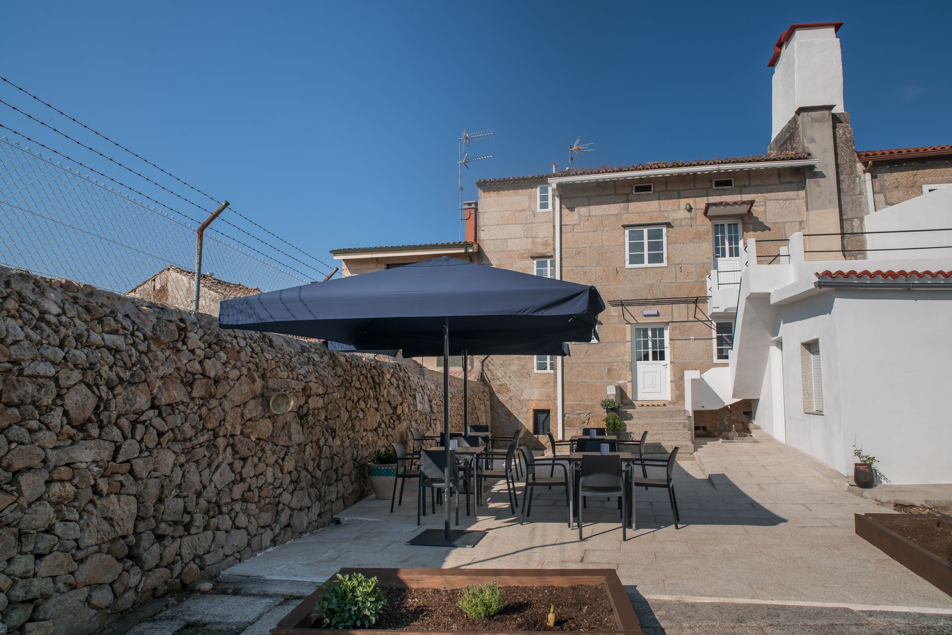 Patio de piedra con mesa, sillas y sombrilla azul junto a un edificio rústico bajo un cielo despejado.