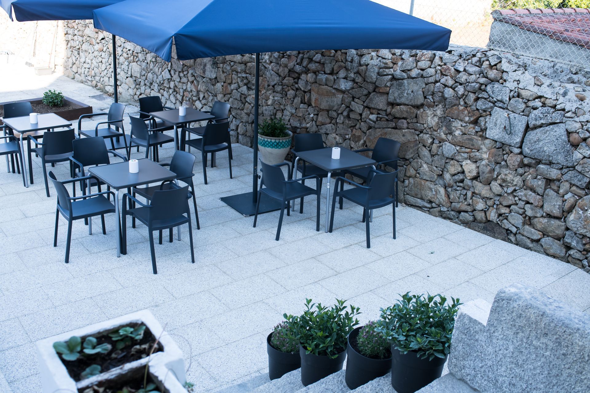 Terraza de cafetería con sillas negras y sombrillas azules junto a un muro de piedra.