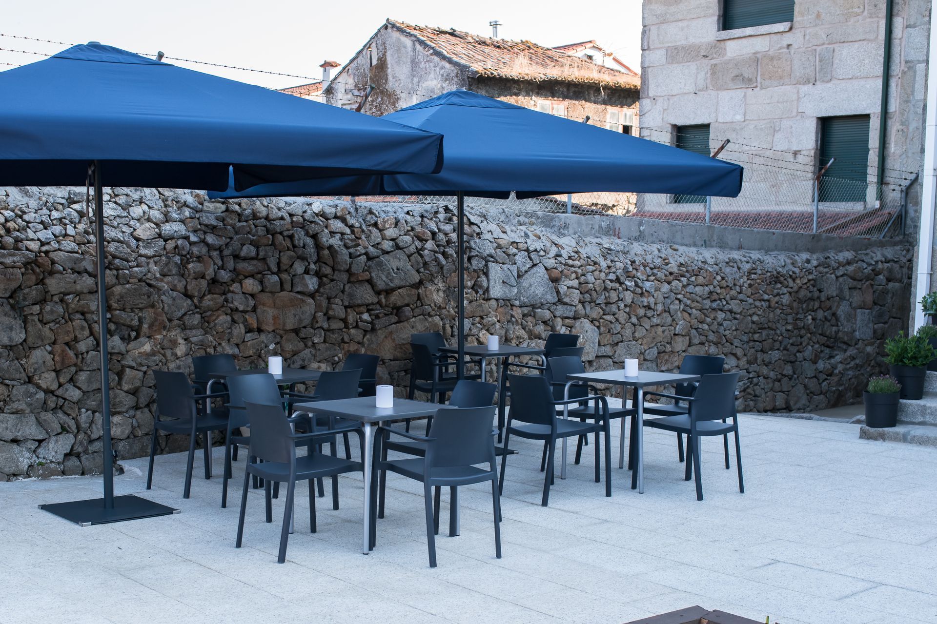 Terraza de café con mesas al aire libre, sombrillas azules y sillas negras en un patio de piedra junto a un muro de piedra.