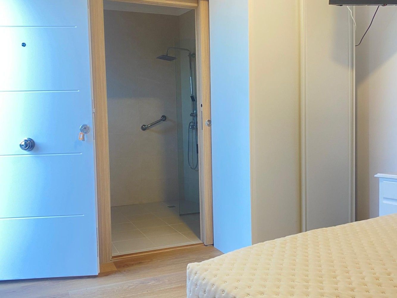 Dormitorio con puerta de armario abierta que da a una zona de ducha o baño, junto a una cama con ropa de cama de color beige.