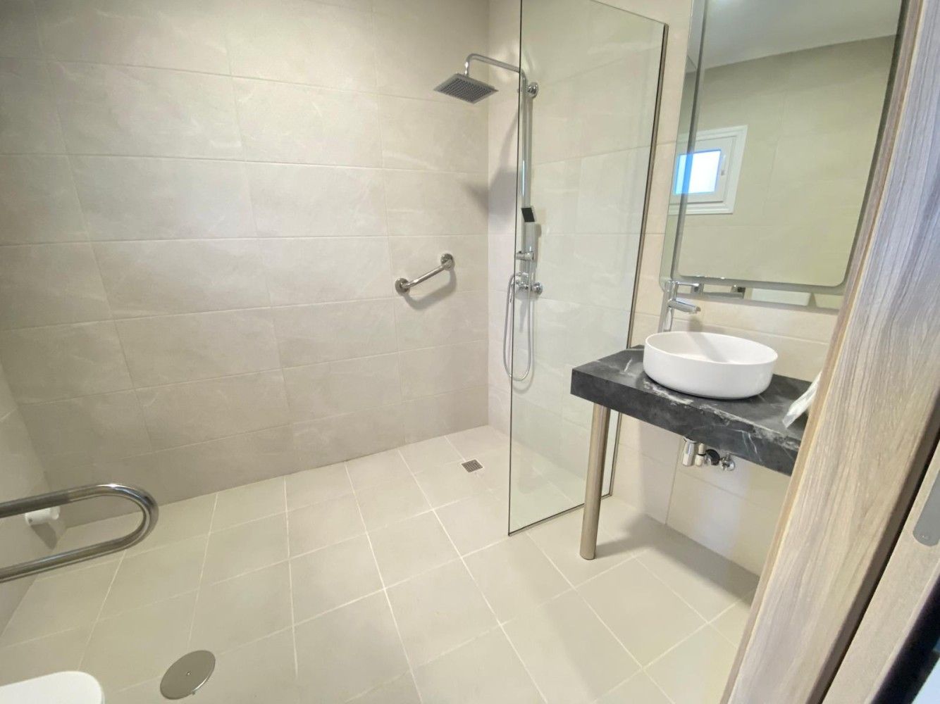 Baño moderno con ducha alicatada, puerta de cristal, asiento de pared, barra de apoyo y lavabo con mueble.