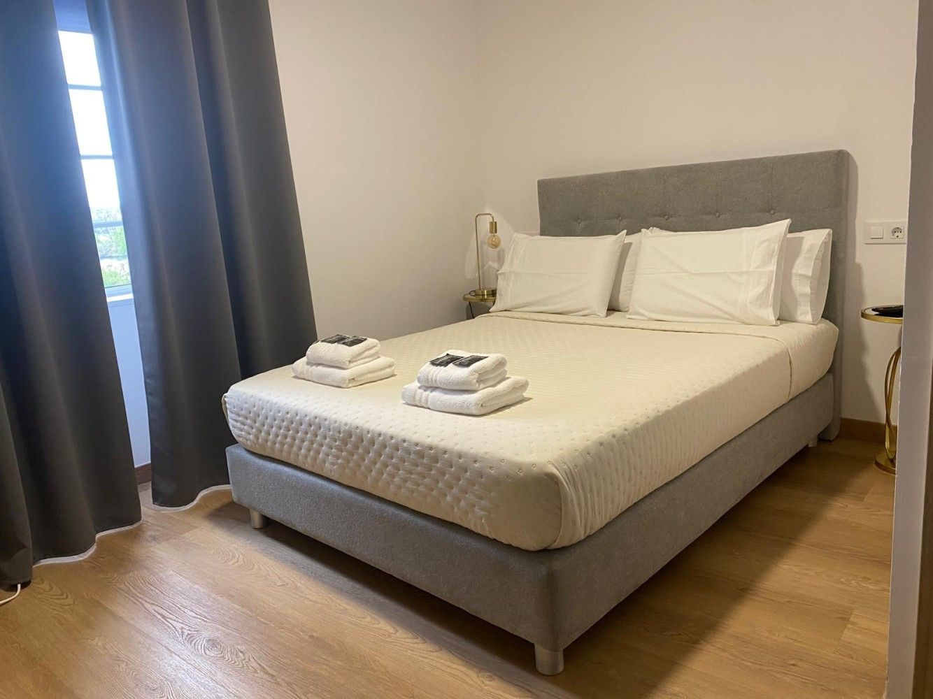 Una cama gris bien hecha con toallas dobladas en un dormitorio luminoso y minimalista con suelo de madera.