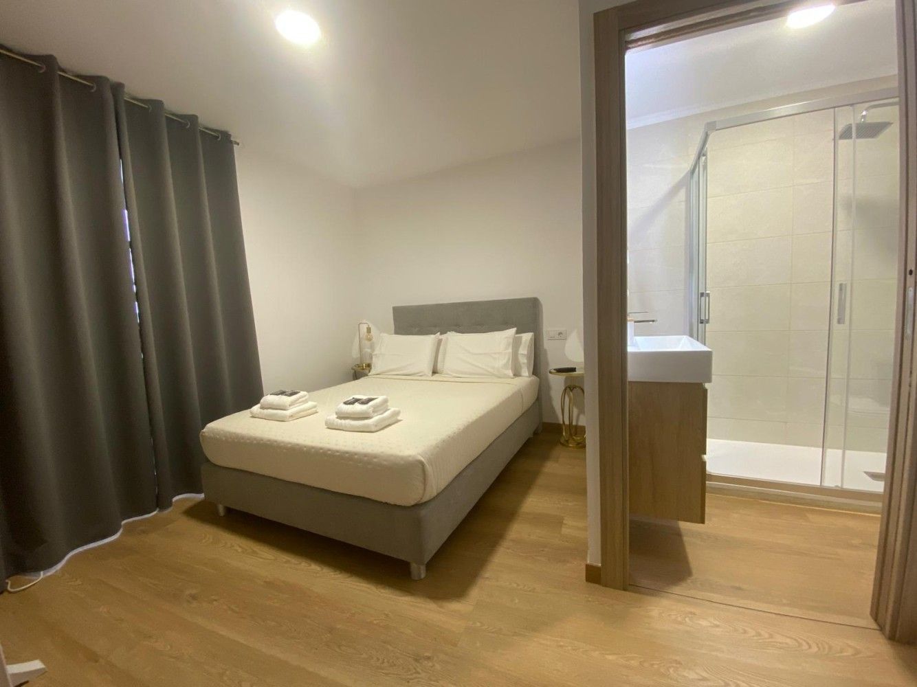 Habitación moderna con cama, cortinas oscuras y baño privado abierto con ducha de cristal.