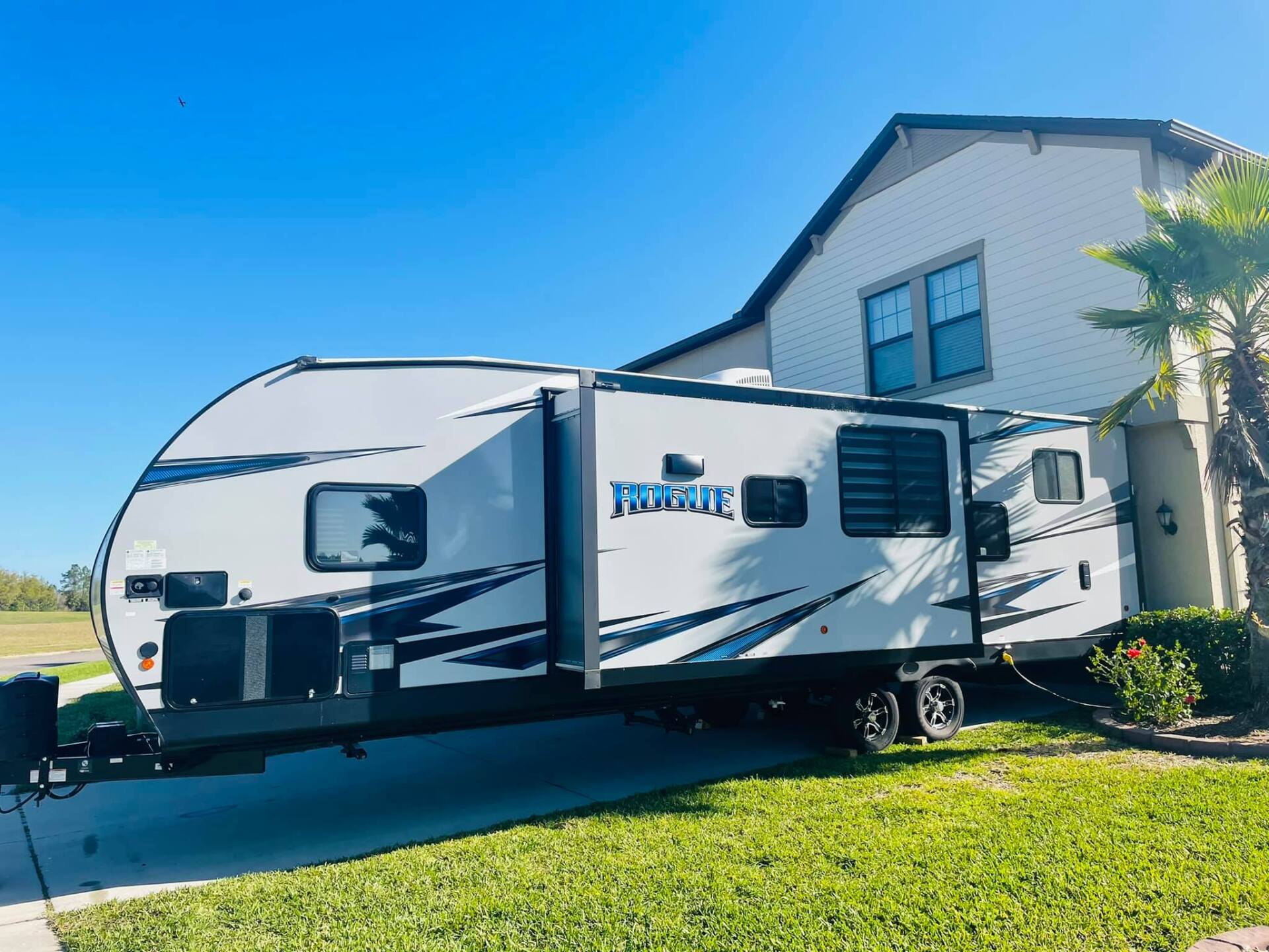 White travel trailer parked beside a beige house on a sunny green lawn under a blue sky