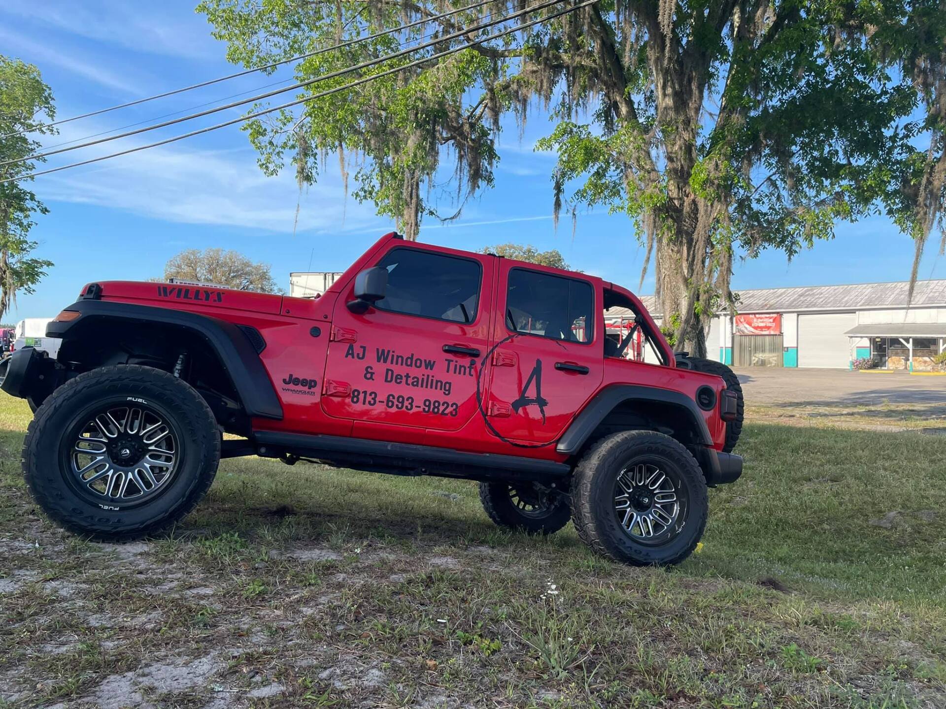 Red lifted Jeep with black wheels parked on grass under trees in a sunny field.