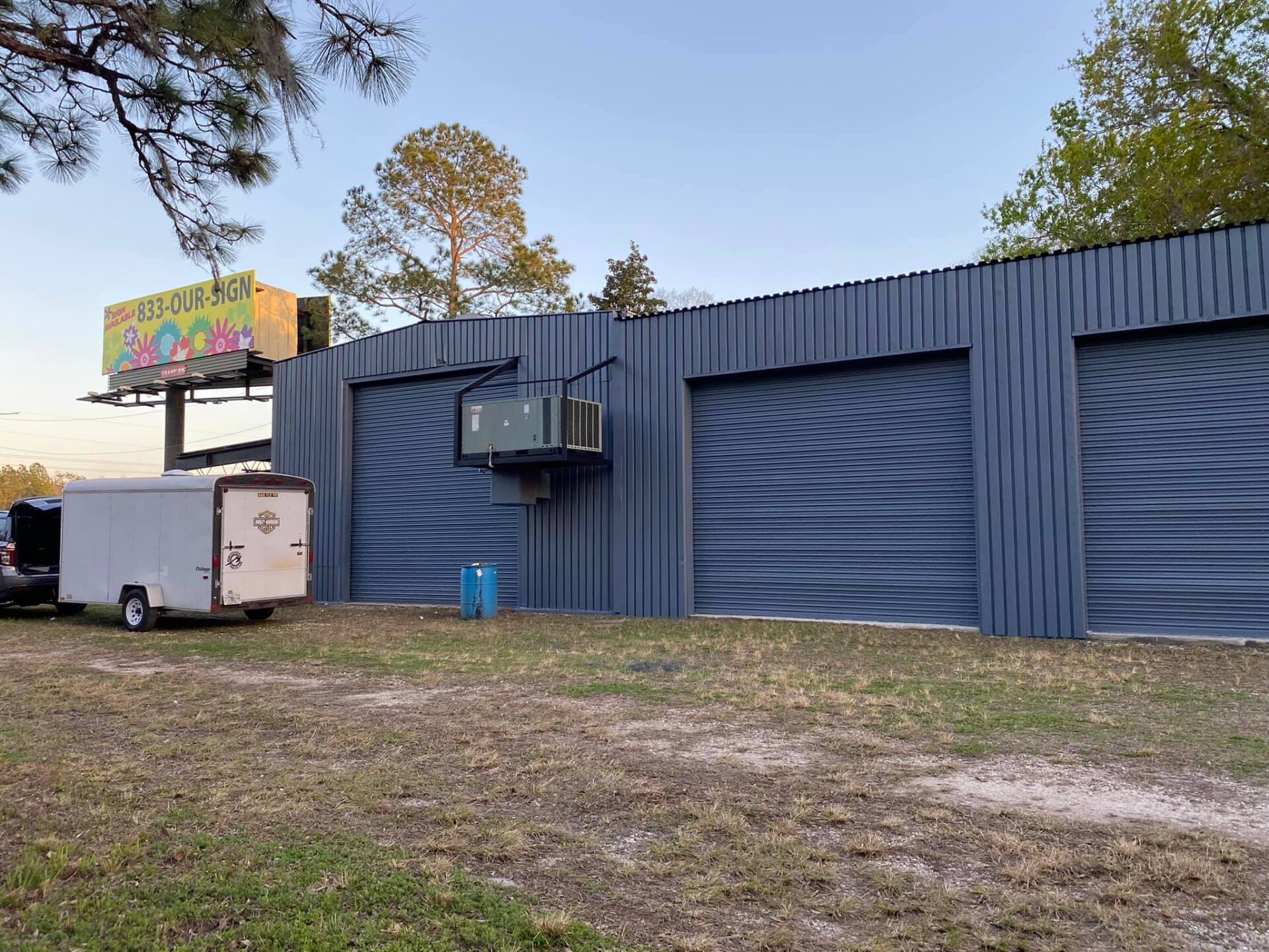 Blue corrugated building with a food truck sign, trailer, and air-conditioning unit on a grassy lot at dusk