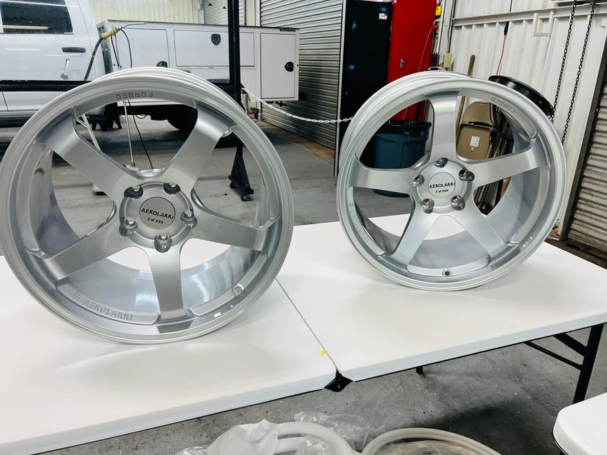 Two silver alloy wheels on white tables in a workshop, one larger rim in front.