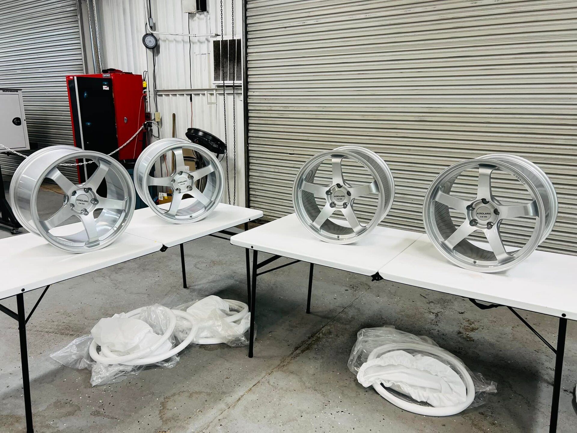 Several silver alloy wheels on folding tables in a workshop, with red equipment in the background