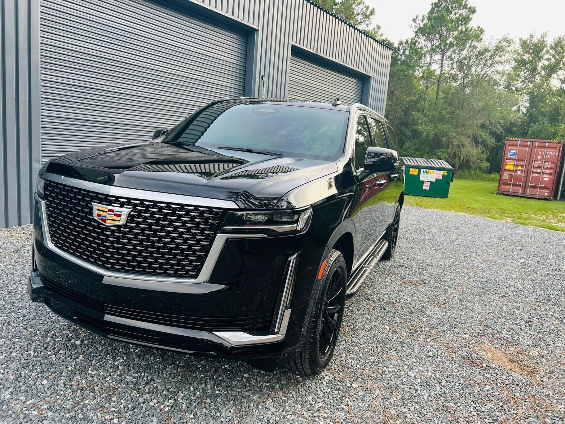 Black Cadillac SUV parked on a gravel driveway beside a garage and dumpster bins.