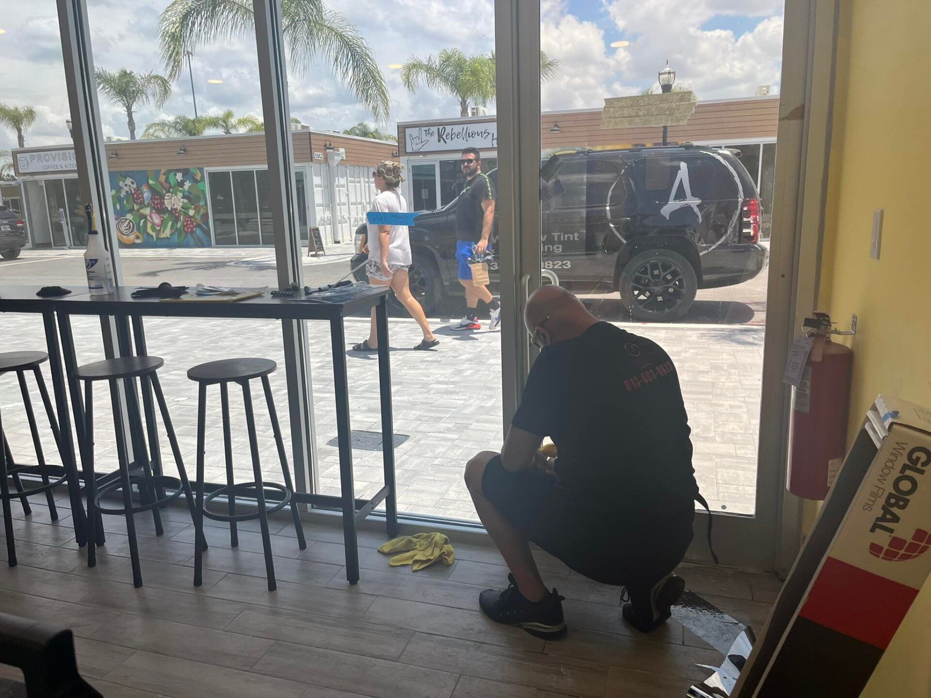 Person crouching by a glass door inside a café, with outdoor street view and stools nearby.