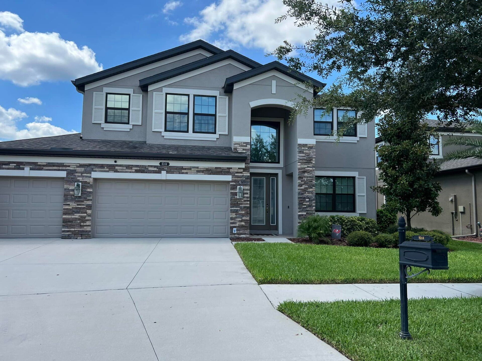 Two-story gray suburban house with a three-car garage, driveway, and front lawn under a blue sky