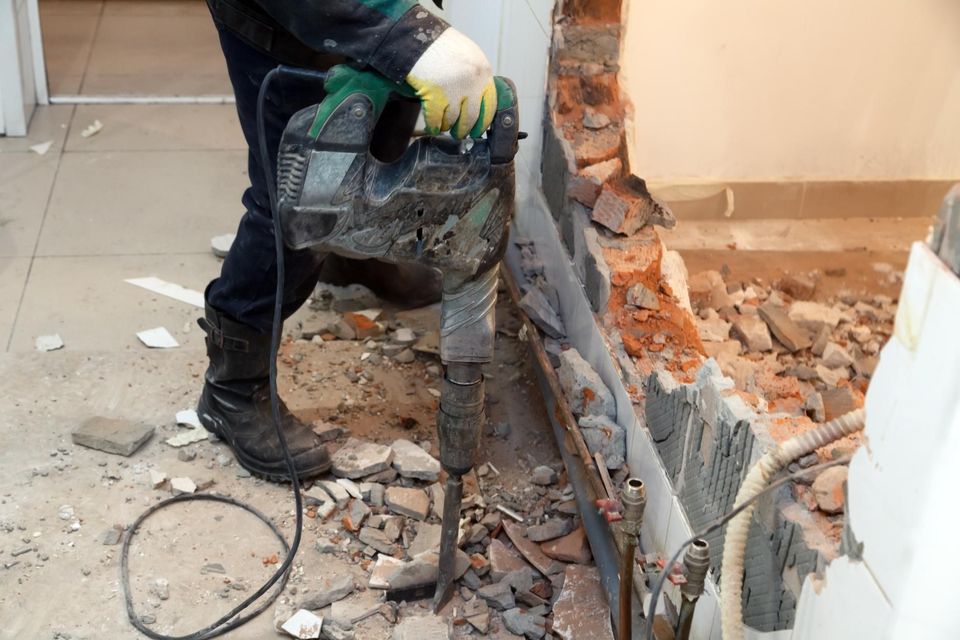 Worker using a jackhammer to break a wall, with rubble scattered on the floor.