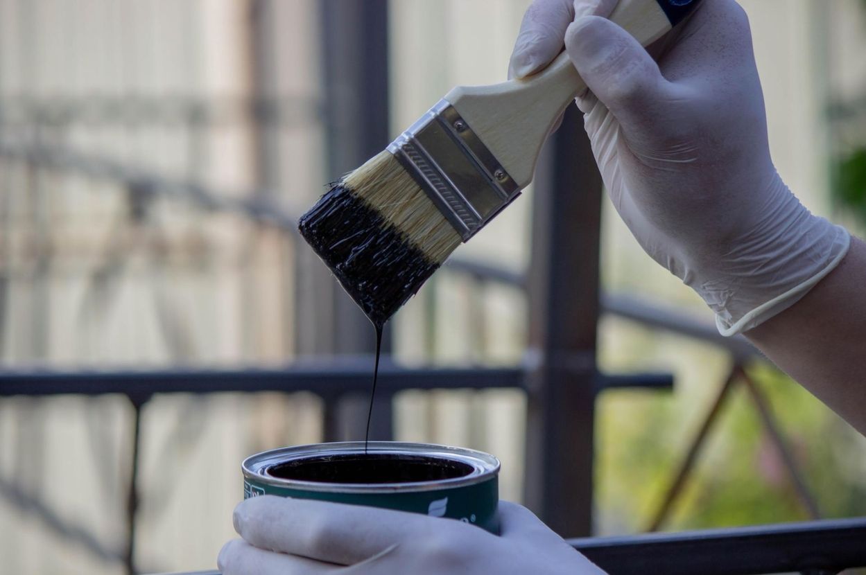 Gloved hand dipping a paintbrush into a small can of dark paint outdoors