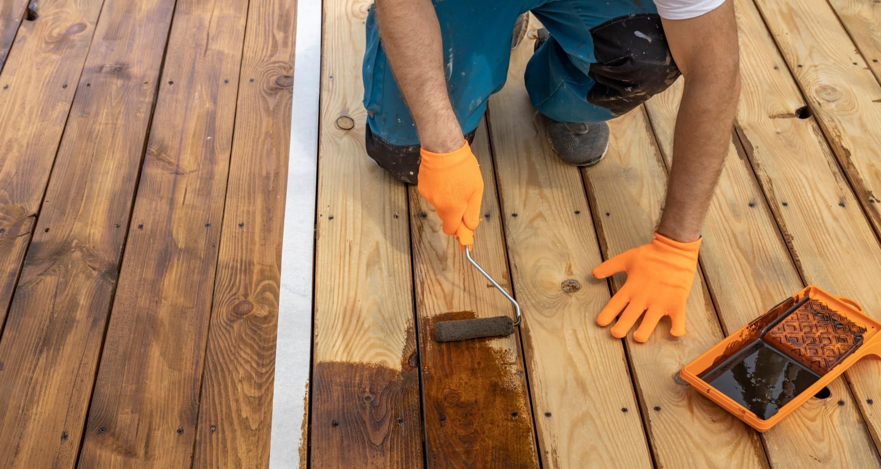 Person staining wooden deck boards with a paint roller, orange gloves, and a tray of stain nearby.