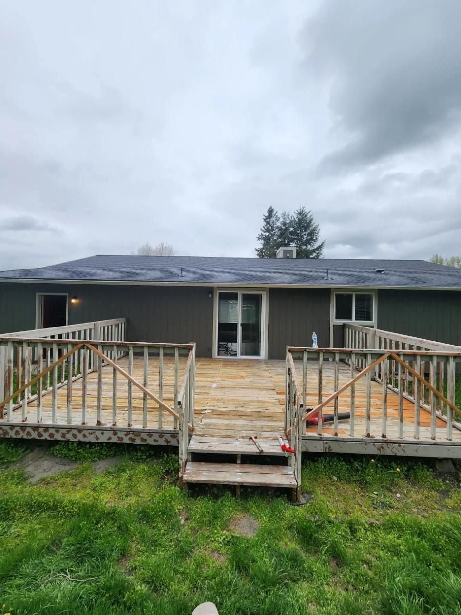 Weathered house deck with broken railing and steps, viewed from a grassy yard under cloudy skies