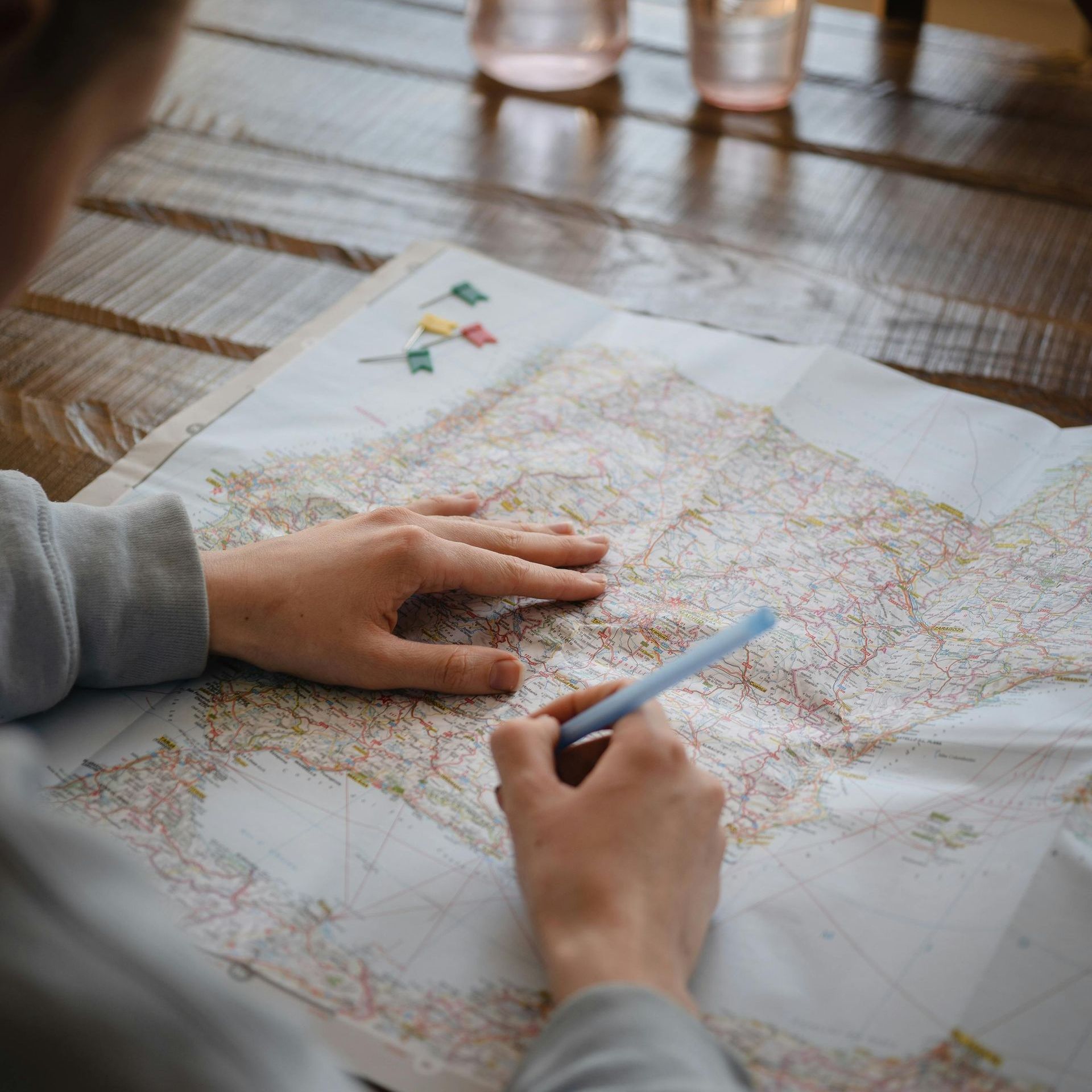 Hands using a pen to mark a map on a wooden table. Small pins are placed on the map.