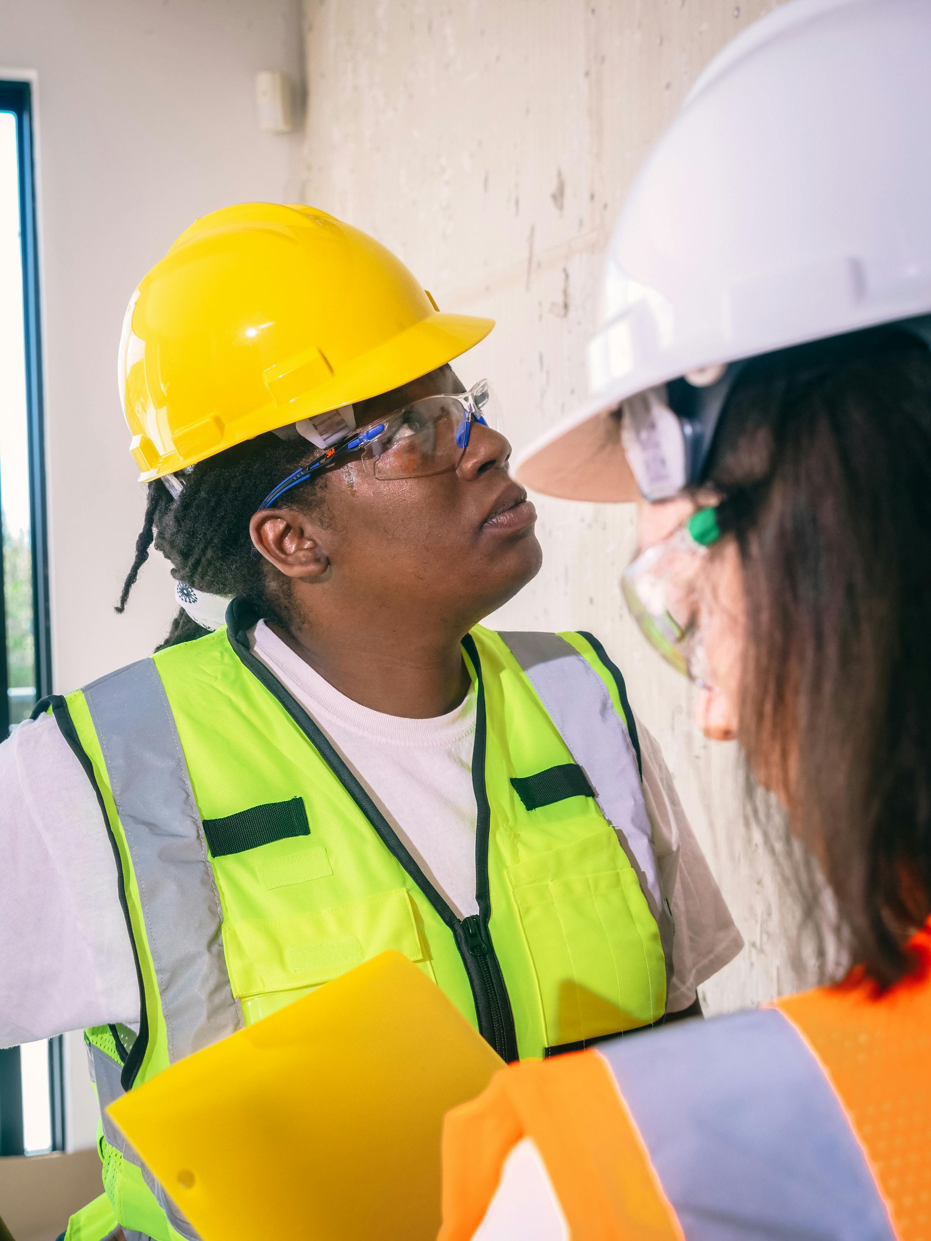 Two safety Inspectors in safety gear talking on site. One in yellow hard hat, holding a clipboard. 