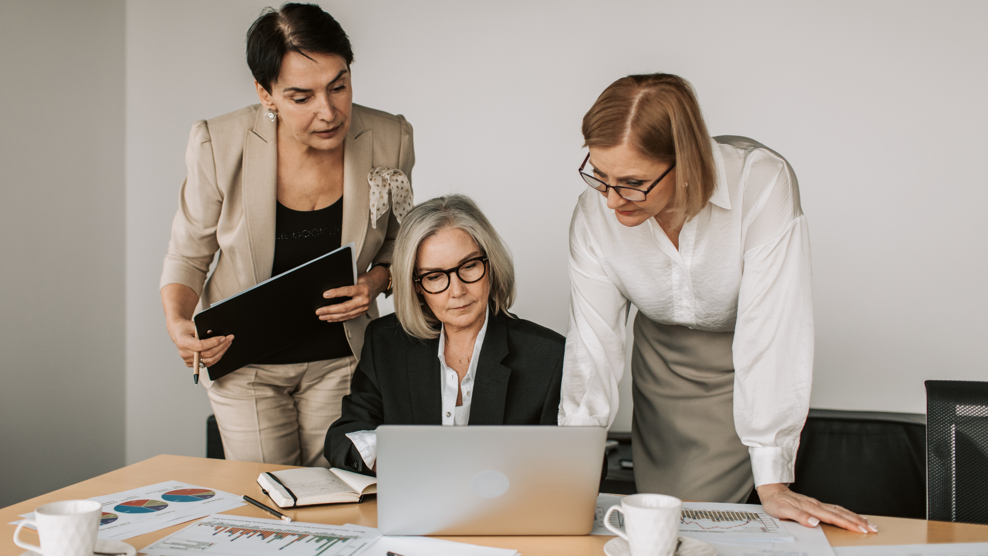 Three women at a table looking at a laptop. One is typing, two are observing. White walls, daytime.