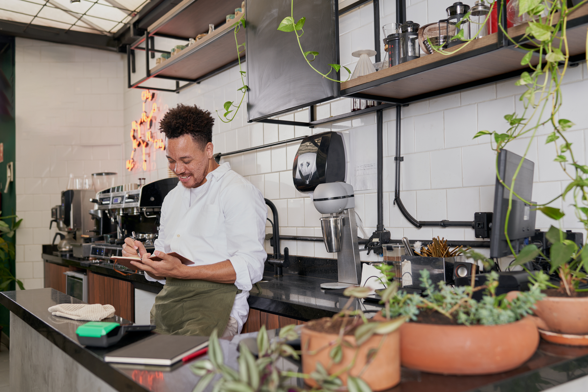 Chef in a modern kitchen writing notes beside a counter with herbs and cooking tools.