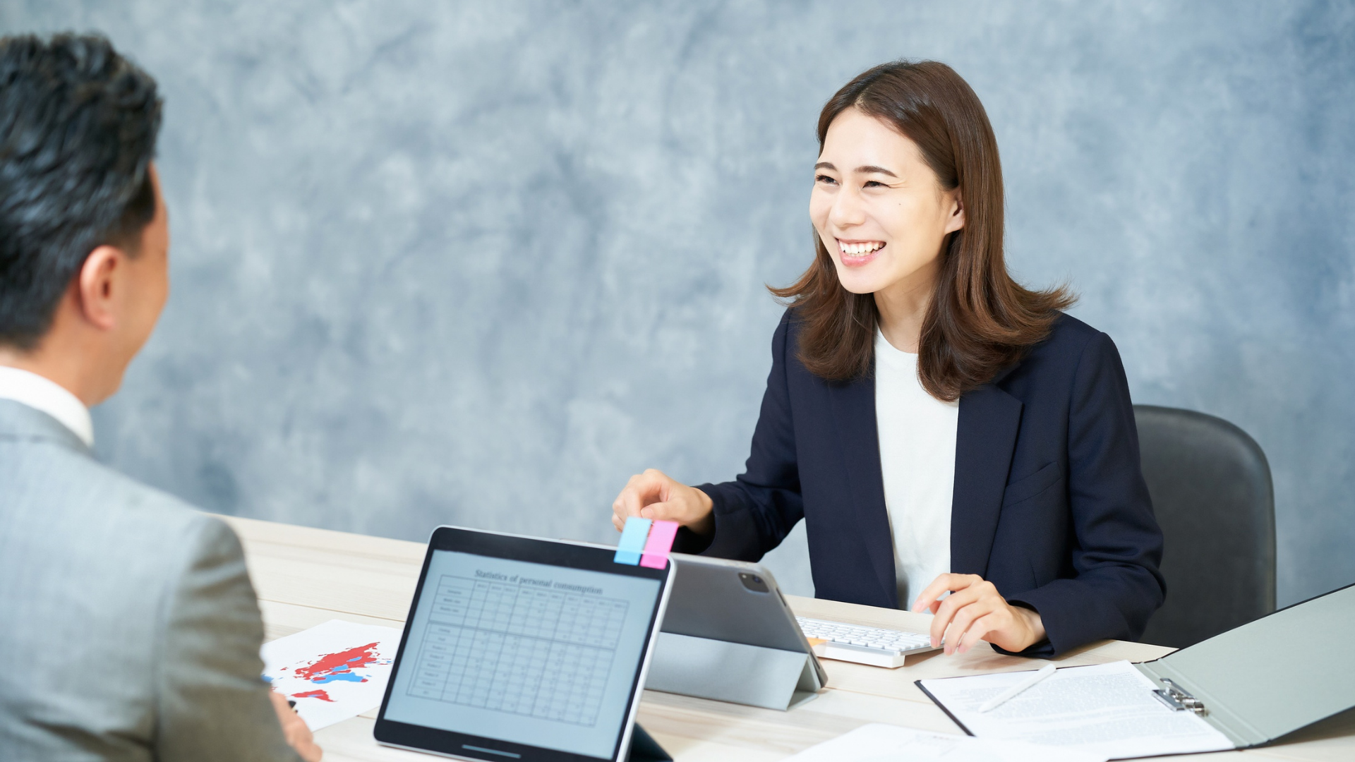 Woman in a blazer smiles, speaking to a man at a desk with a tablet and papers. Gray wall background.