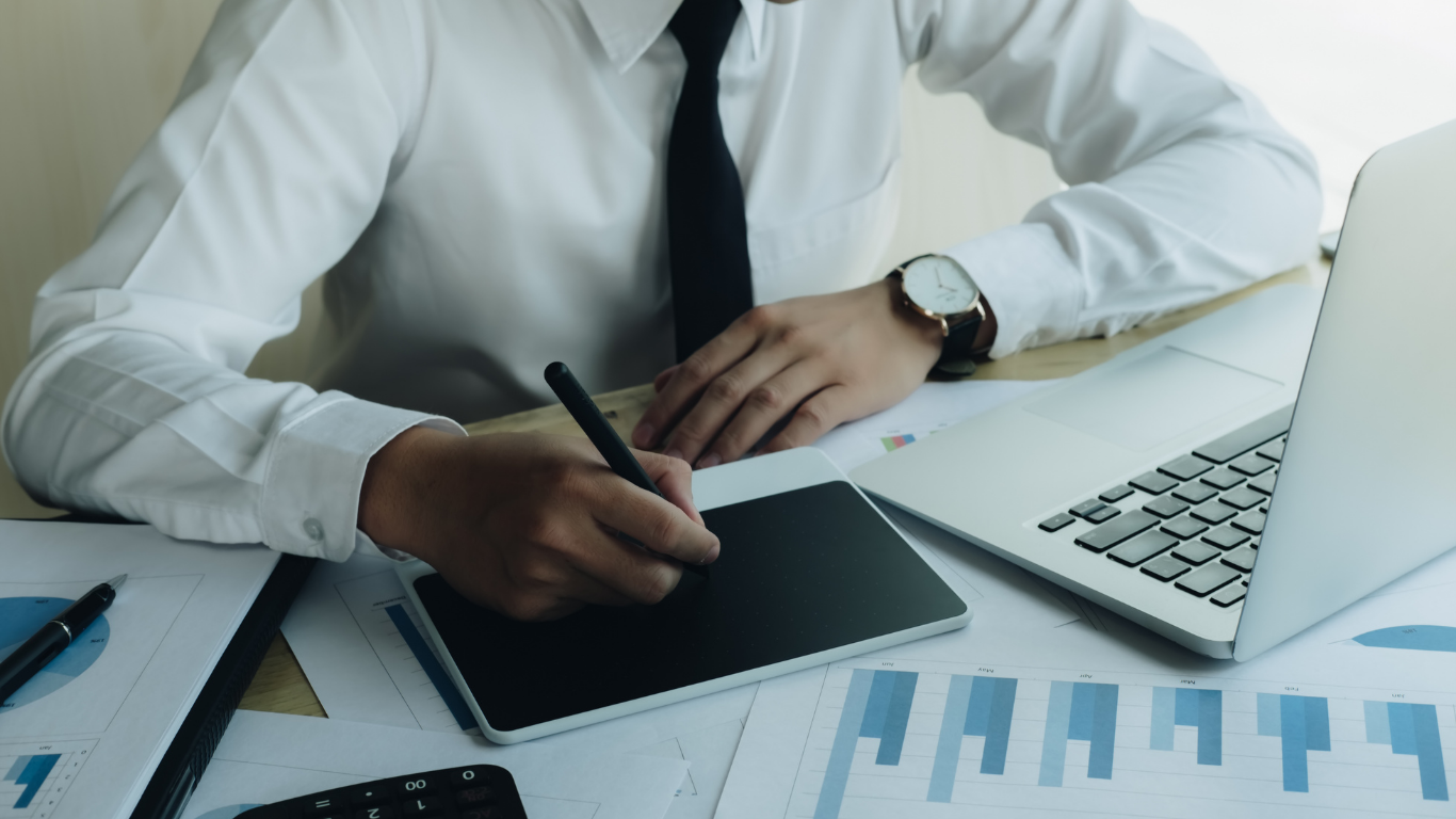 Person in a white shirt and black tie writing on a tablet at a desk with laptop and charts.