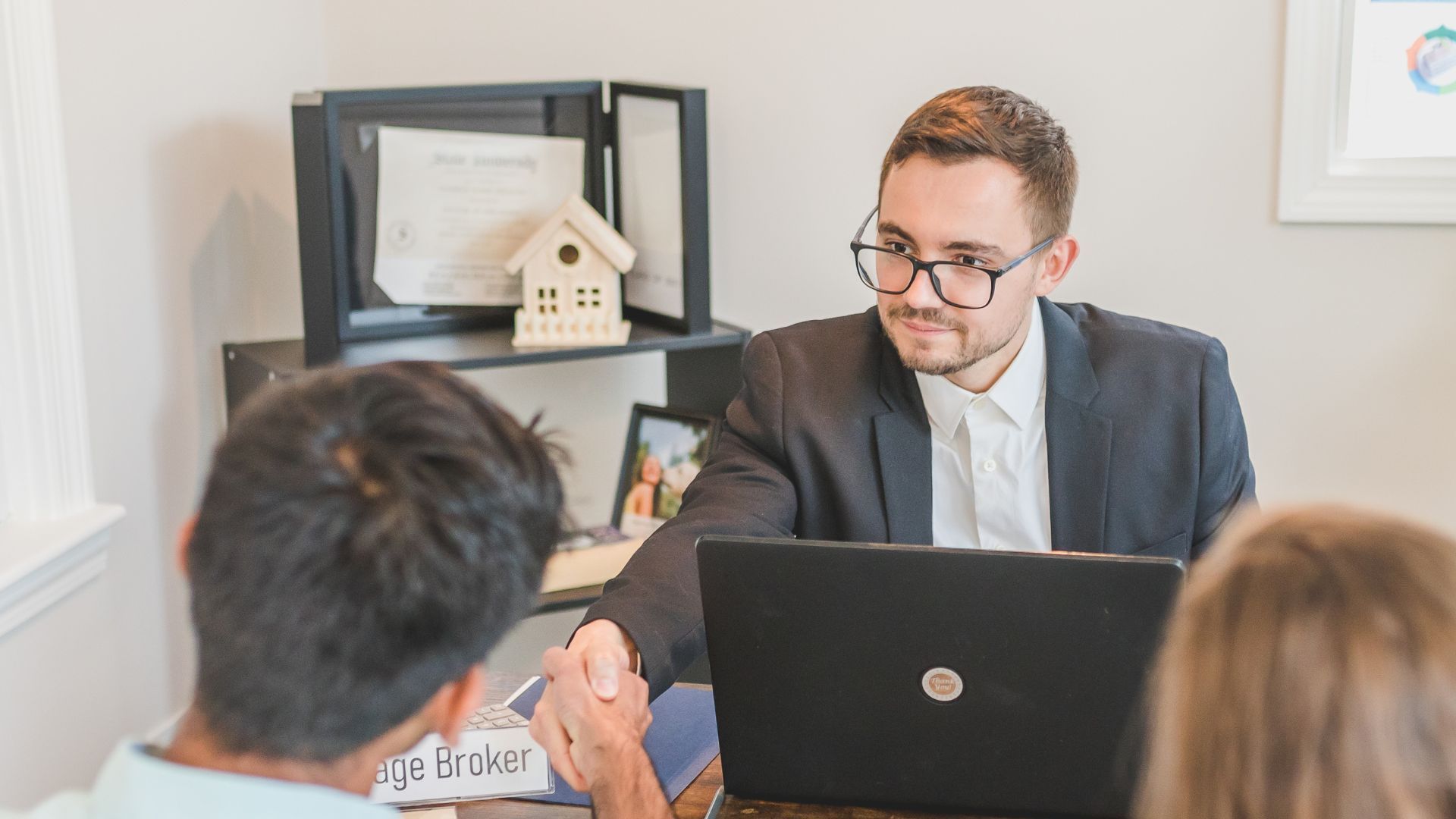 Meeting in an office with a man in glasses using a laptop while talking to two seated colleagues.