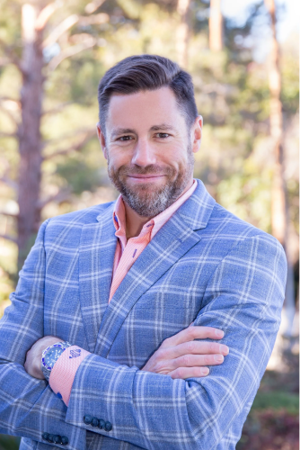 A man in a blue suit and pink shirt is standing with his arms crossed.