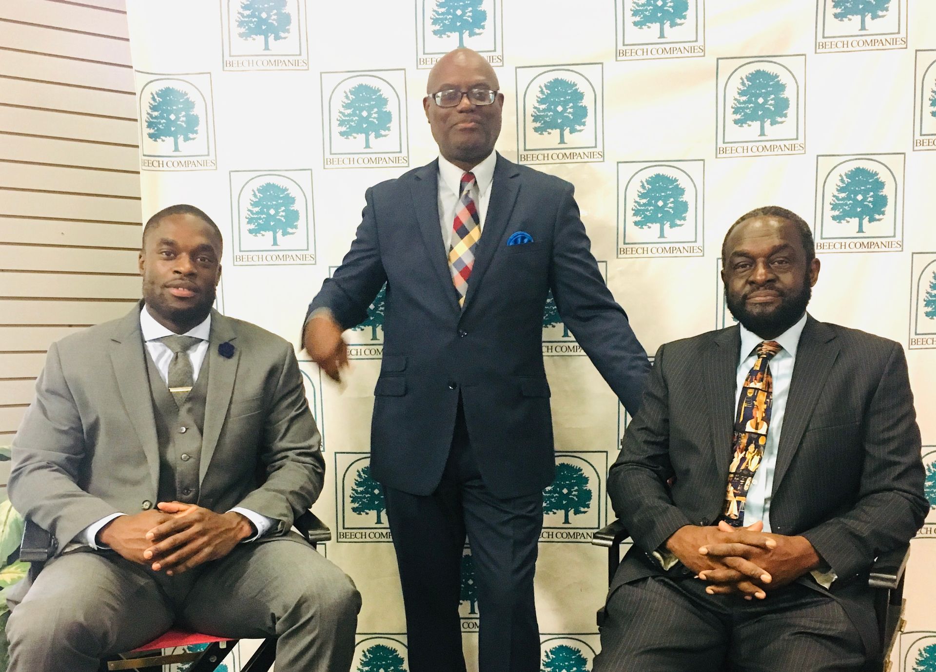 Three men in suits pose in front of a logo backdrop; one stands, two sit.