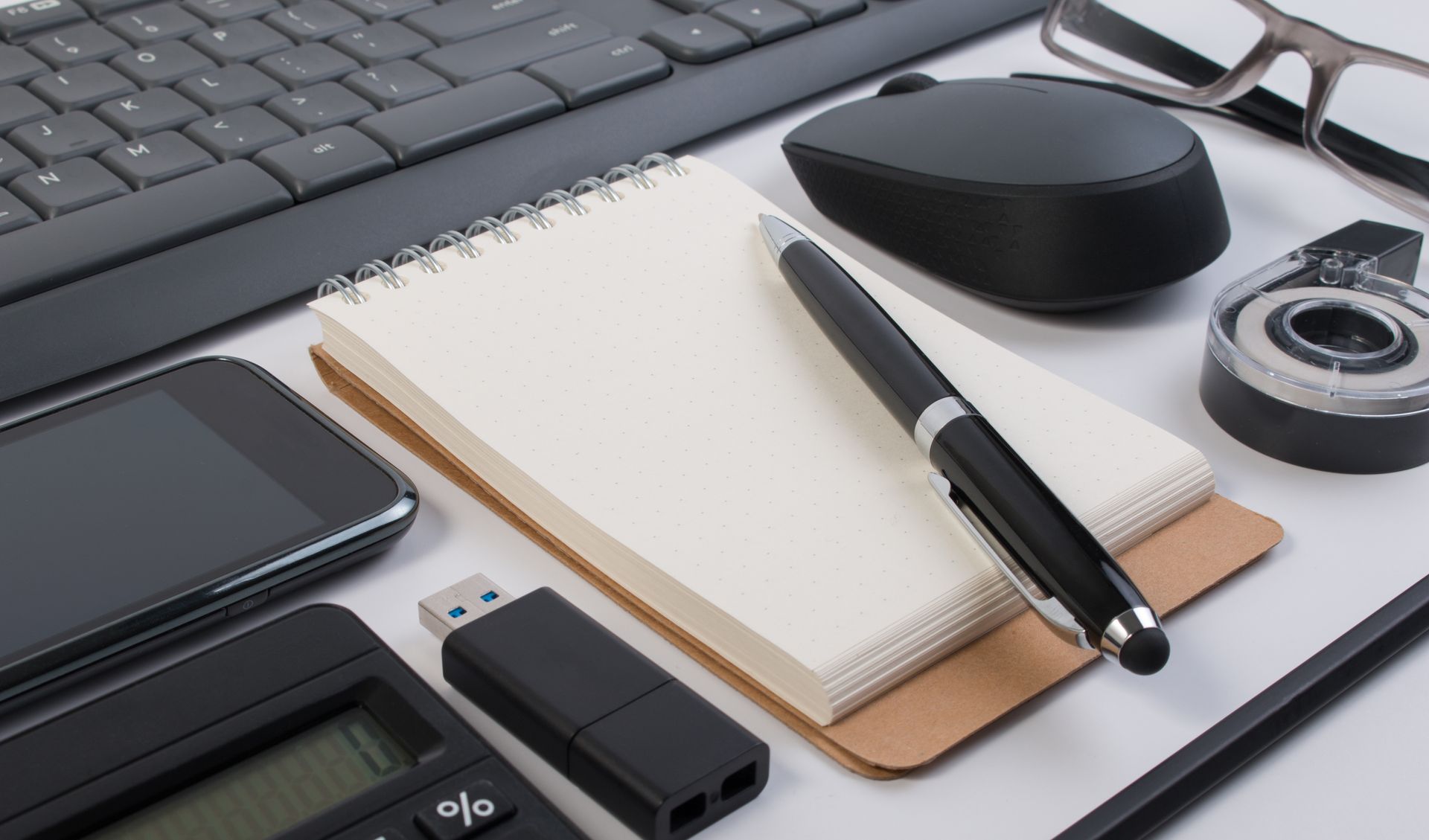 Office desk setup with keyboard, mouse, notepad, pen, phone, calculator, glasses, and USB drive.