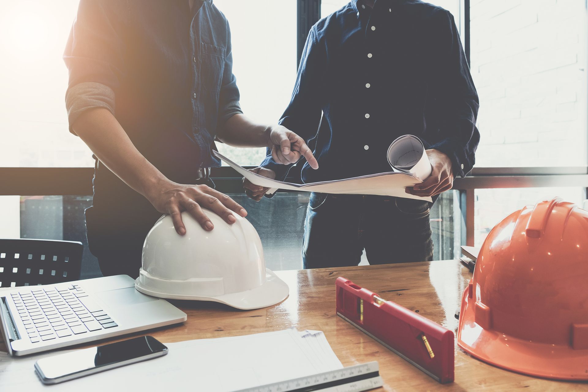 Two people reviewing blueprints at a desk with hard hats, a laptop, and tools.