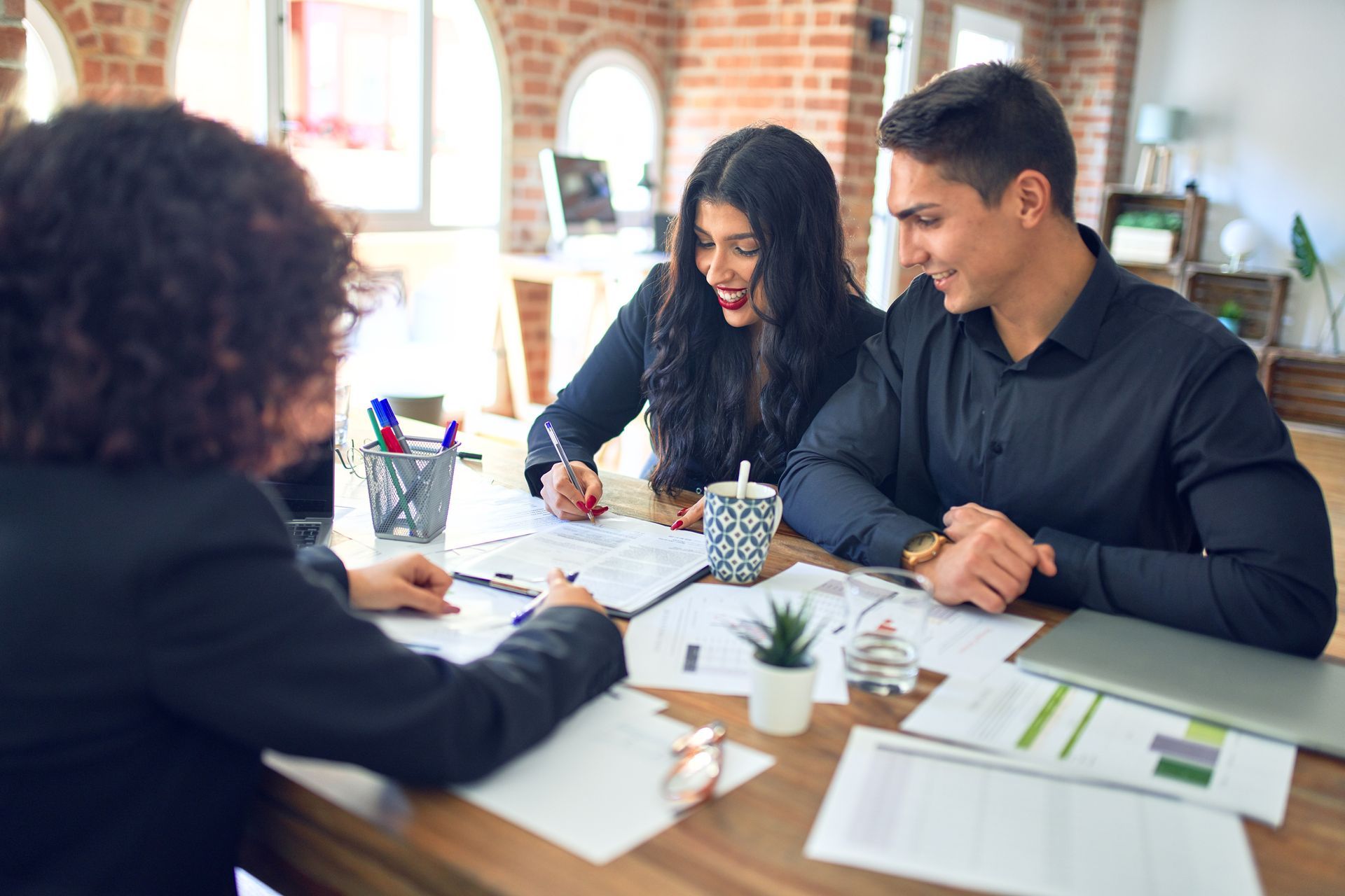 Woman signs document with a couple; discussing paperwork in an office.