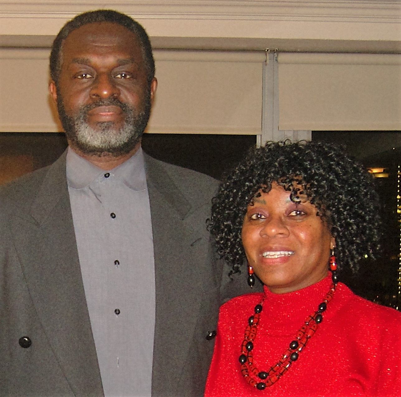 Man and woman smiling, standing indoors; man in suit, woman in red dress.