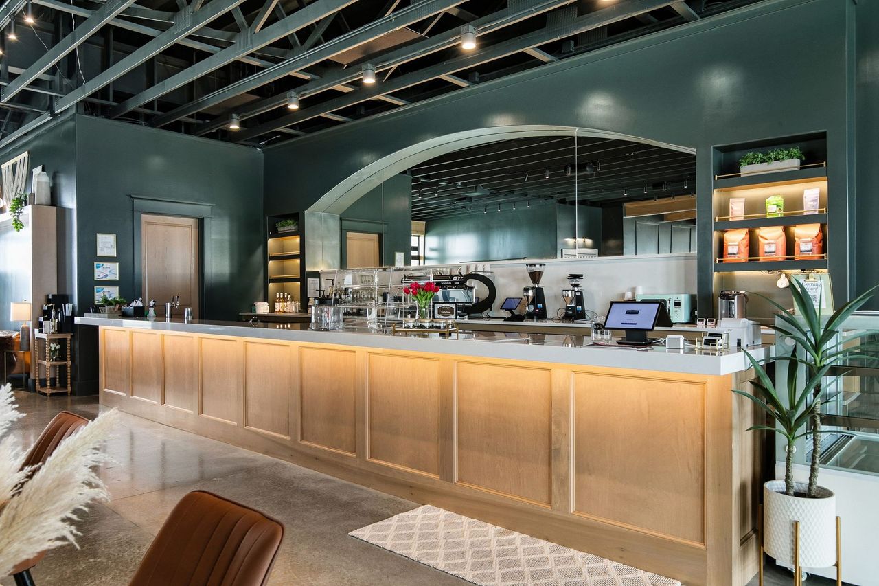 Modern coffee bar with warm wood counter, espresso machines, and dark green interior.