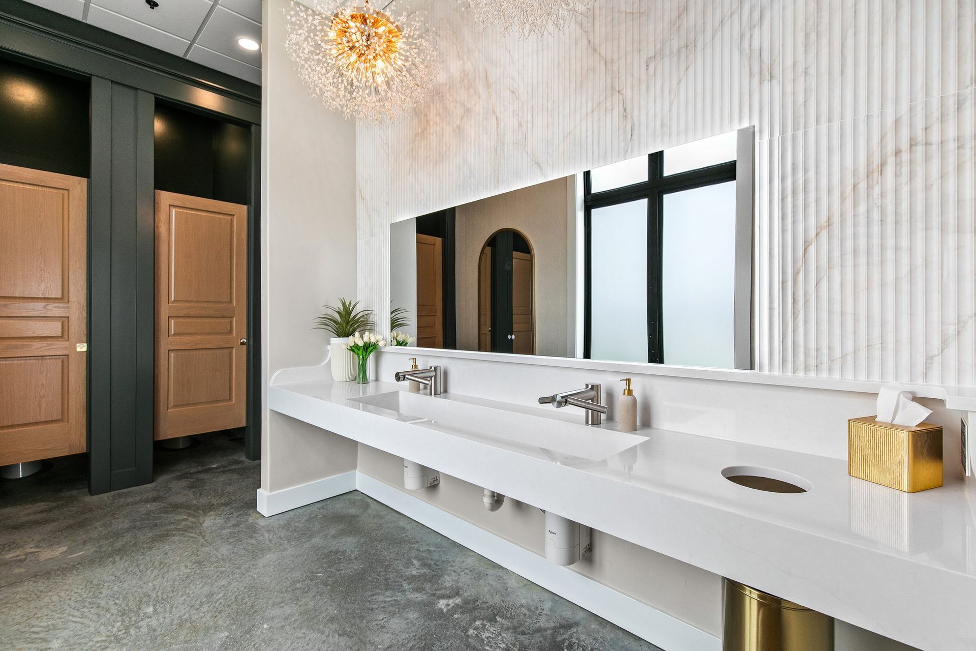 Modern restroom with long white vanity, large mirrors, wooden stalls, and dark stone flooring