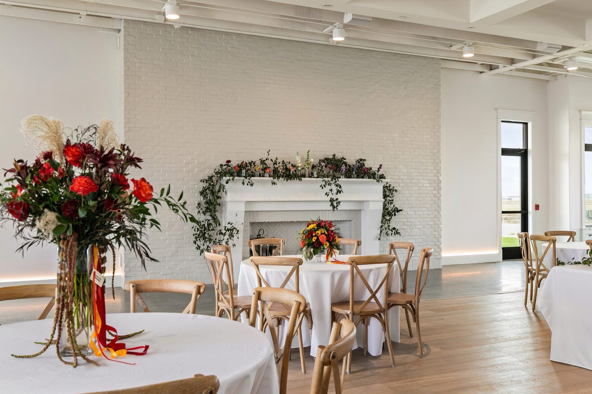 Elegant event hall with white tables, wooden chairs, and floral centerpieces beside a fireplace.