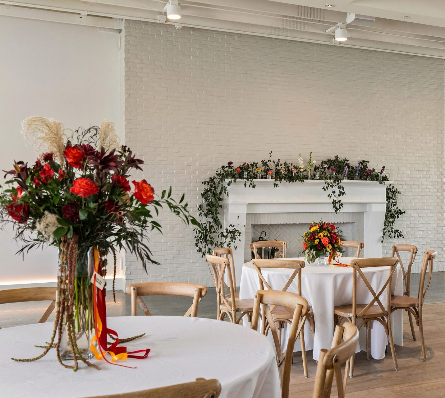 Elegant event room with white tables, wooden chairs, and red floral arrangements by a decorated fireplace