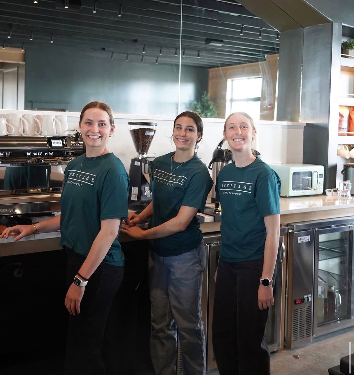 Three café staff in teal shirts standing behind a coffee counter, smiling at the camera.