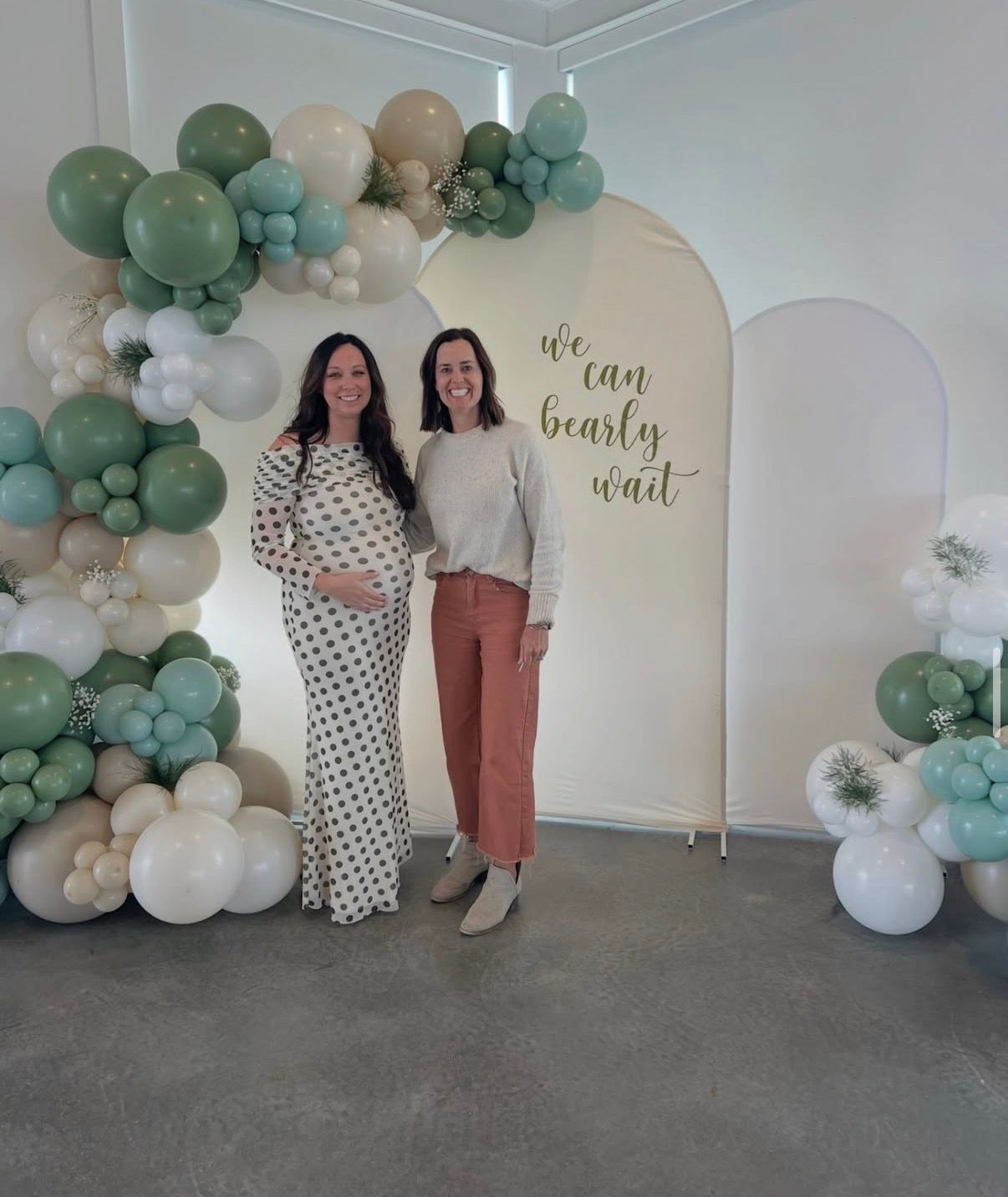 Two women stand in front of a balloon arch and a “Oh baby” backdrop at a baby shower.