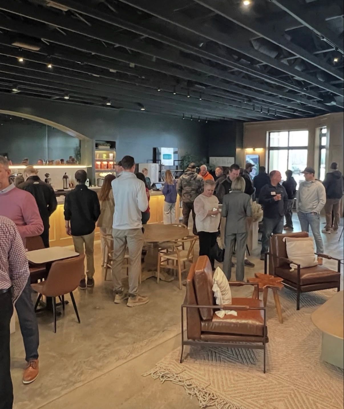 Crowd gathered in a modern lounge or café with wooden chairs and tables under a dark ceiling.
