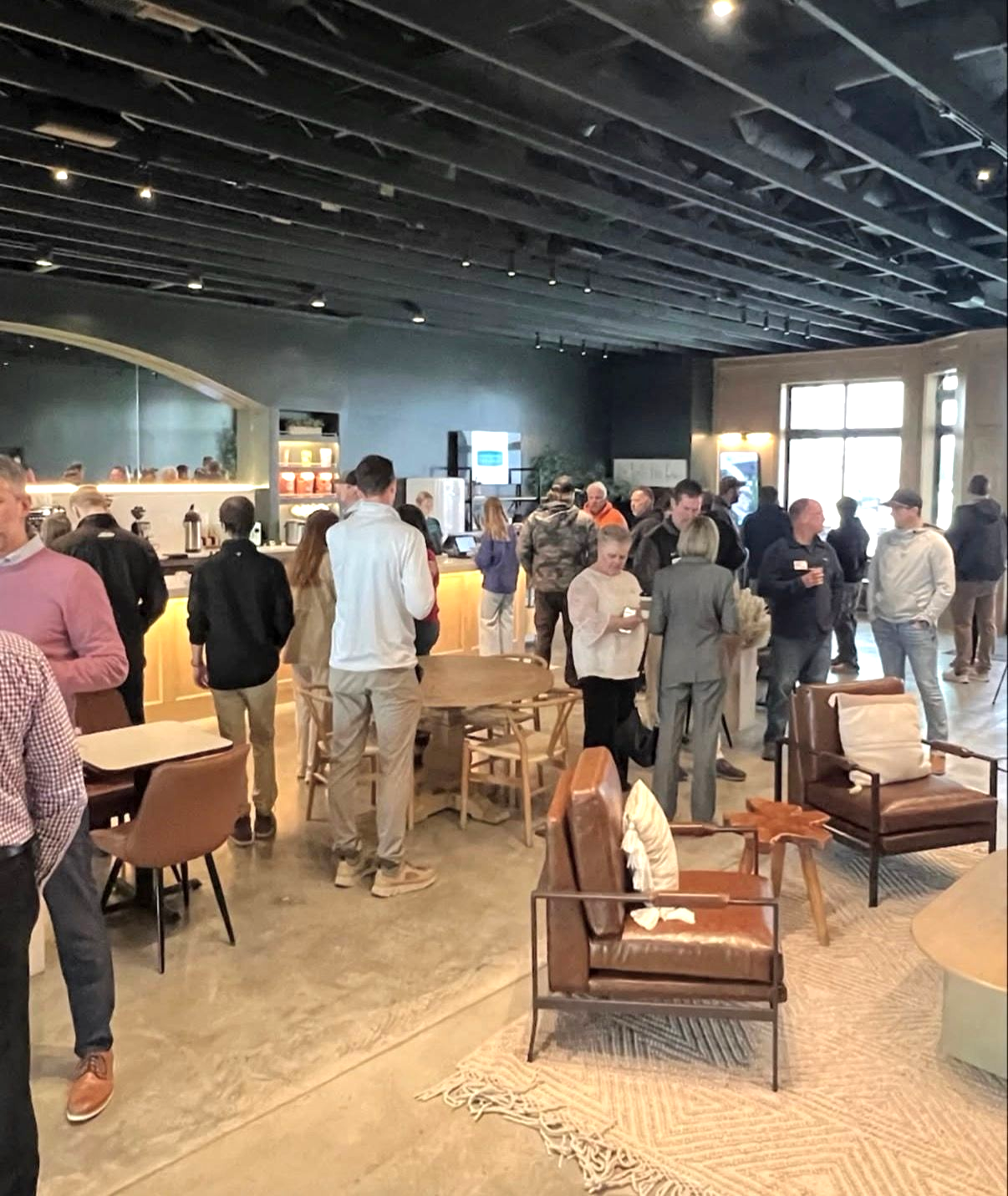 People gathered in a spacious lounge with tables, chairs, and a bar under a dark ceiling.